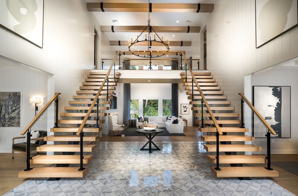 Symmetrical floating wood staircase with glass railings in a modern farmhouse foyer featuring exposed beams, chandelier, and geometric tile flooring